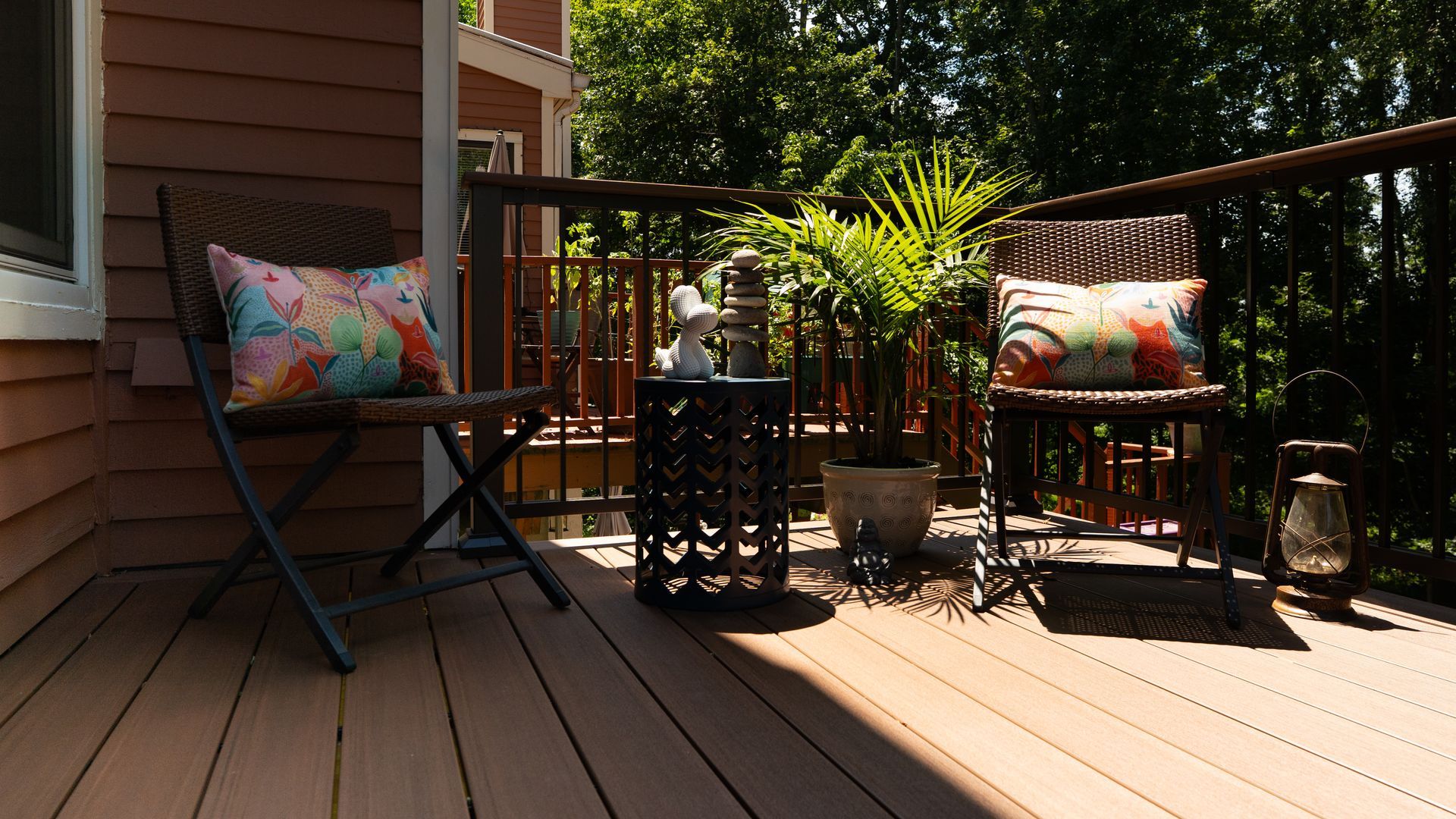 Sunlit wooden deck with two black chairs, colorful pillows, potted plants, and a railing overlooking trees.