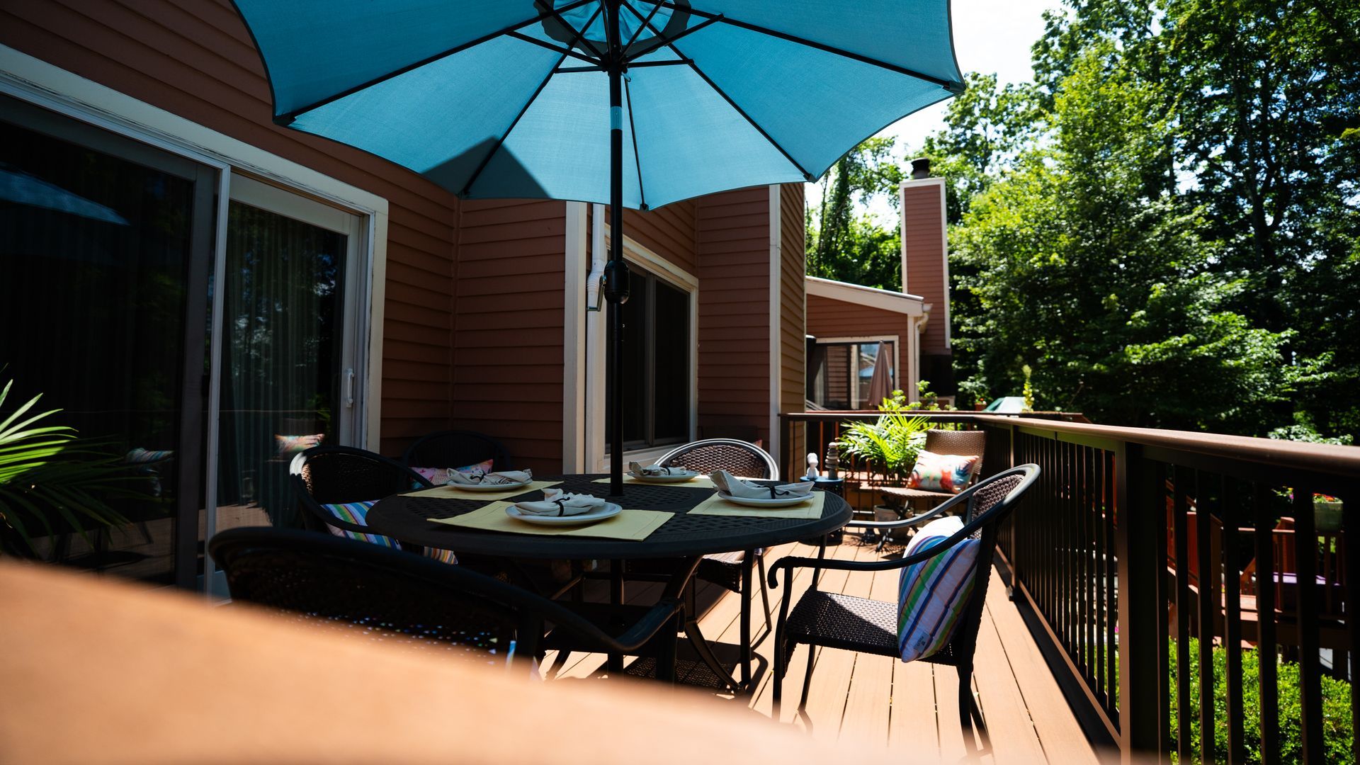Patio deck with table and chairs under a blue umbrella beside a house, surrounded by trees