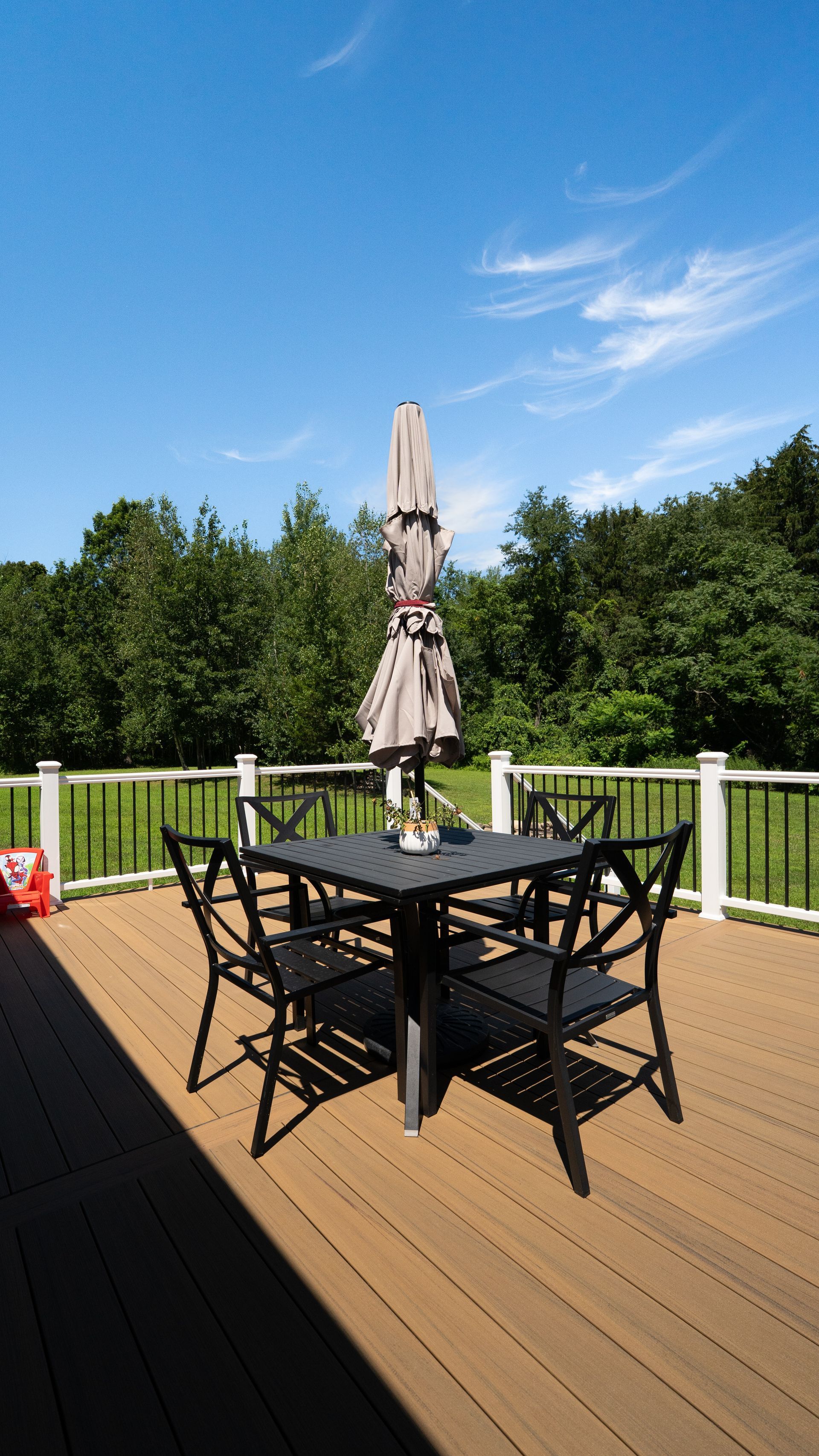 Wooden deck with black patio table, chairs, and closed umbrella overlooking green trees and blue sky