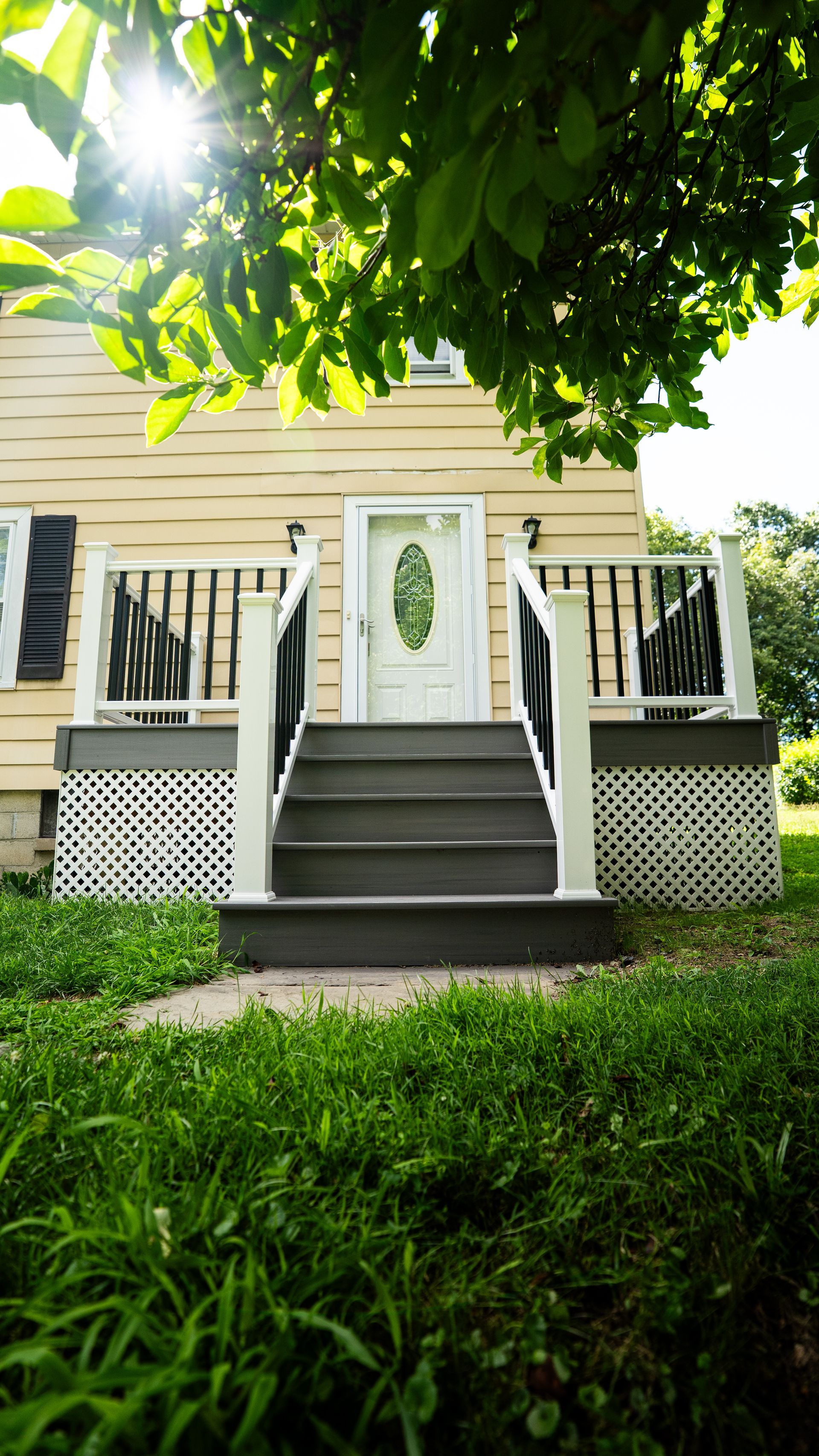 Front porch of a yellow house with black railings, white door, and steps under leafy green trees.