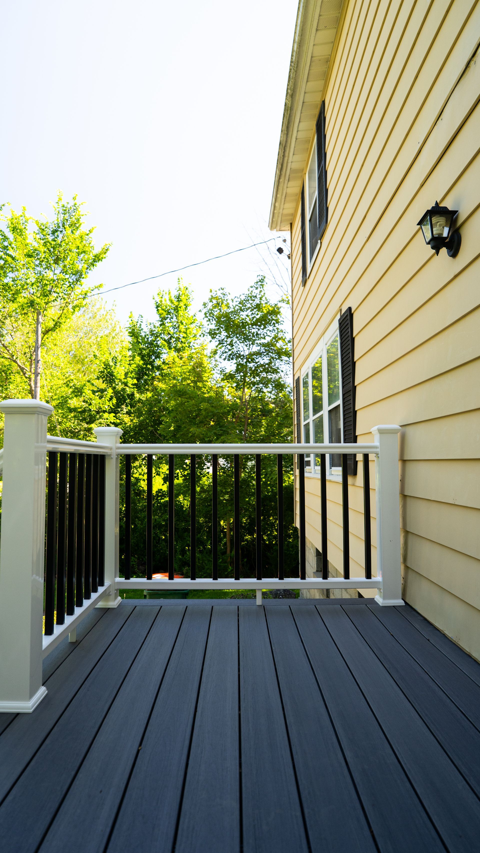 White porch railing beside beige house wall with dark gray deck boards and trees in the background