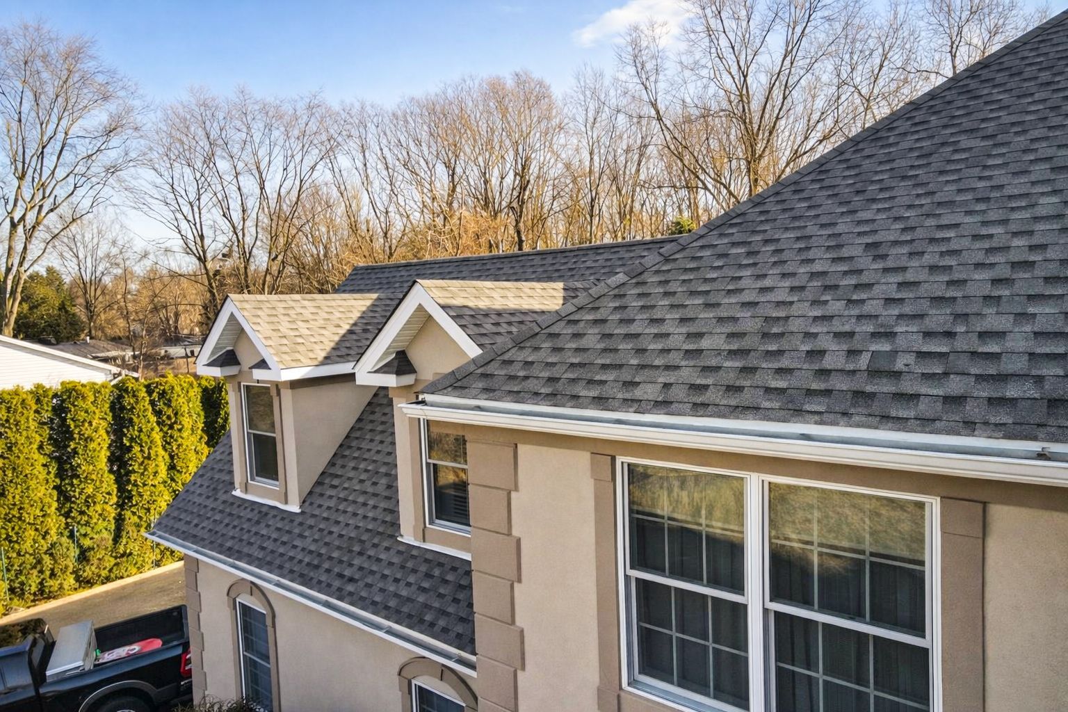 Suburban house roof with gray shingles, dormer windows, and trees in the background