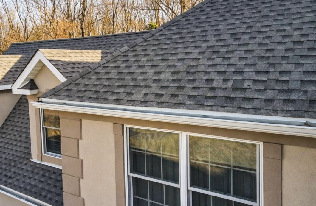 Suburban house roof with gray shingles and beige siding, seen from an angle with windows below
