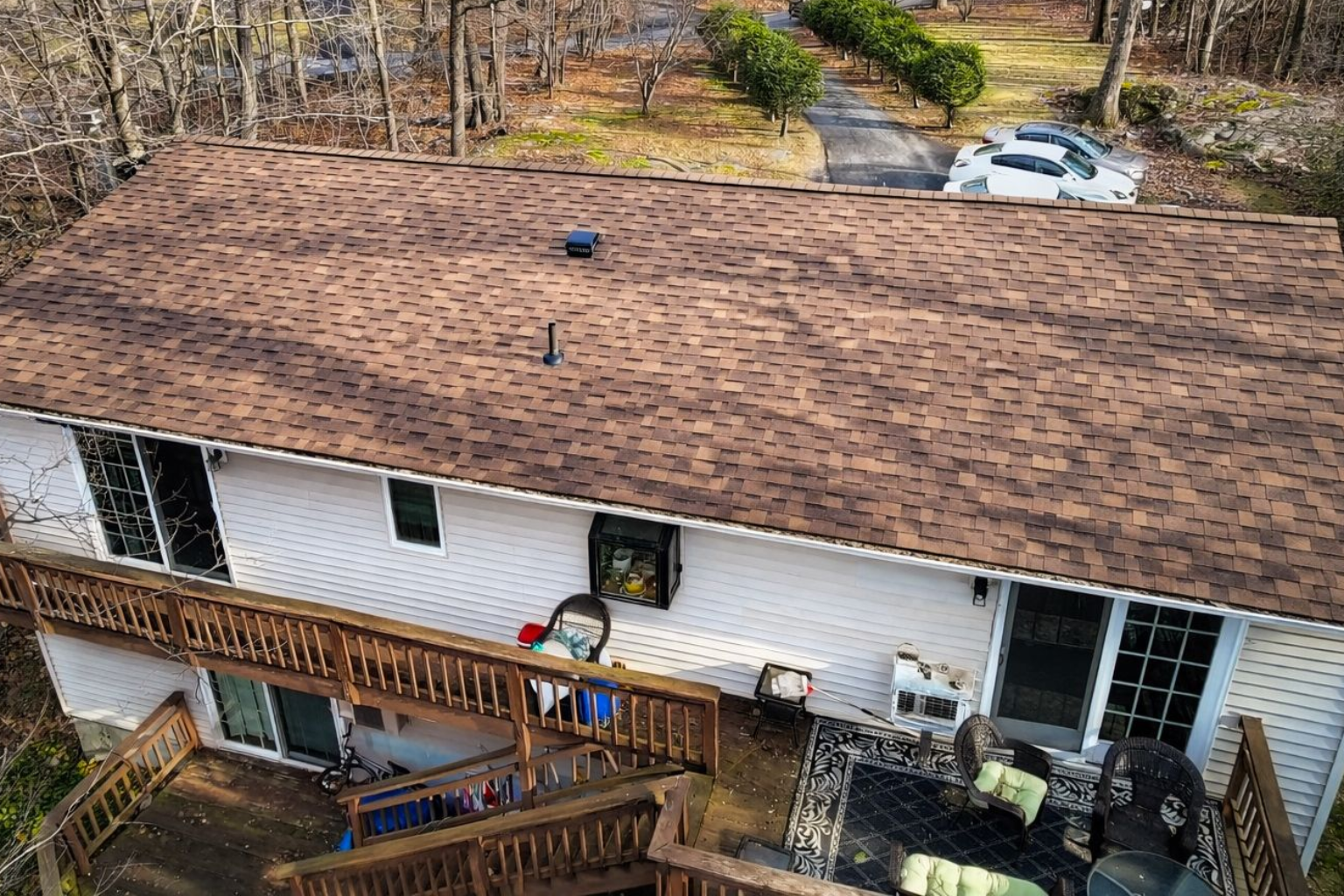 Aerial view of a white house with a brown roof, wooden deck, and wooded yard with a white car nearby
