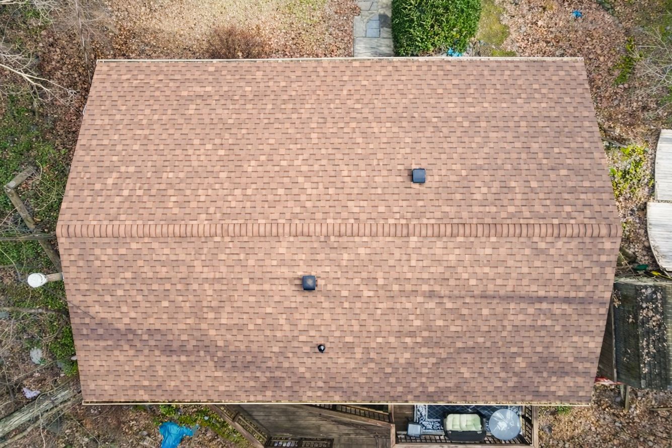 Aerial view of a tan shingled roof with two vents, surrounded by trees and yard debris.