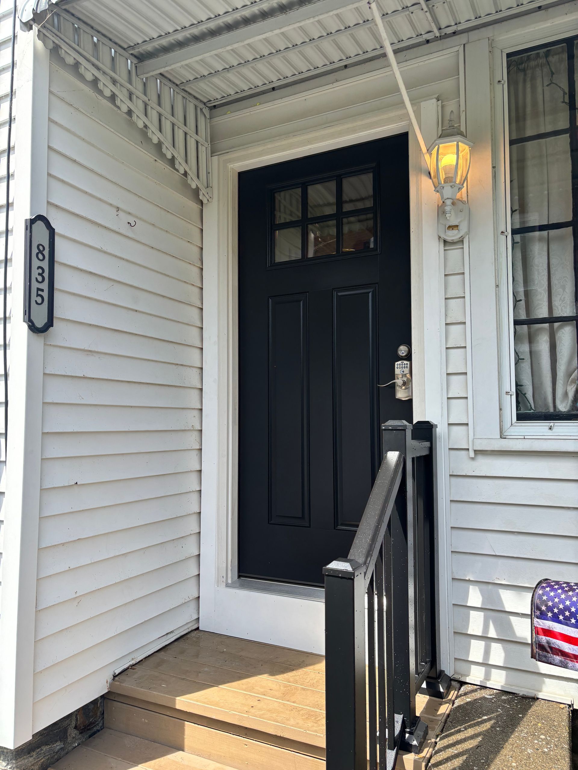 Front porch with black front door, white siding, wall lantern, and steps with black railing.