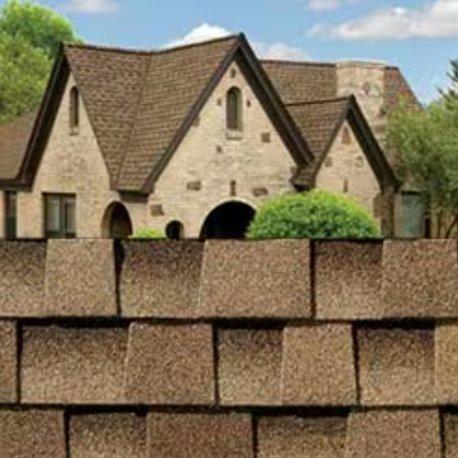 Stone house behind stacked tan cinder blocks, with a brown shingle roof and green bushes in front.