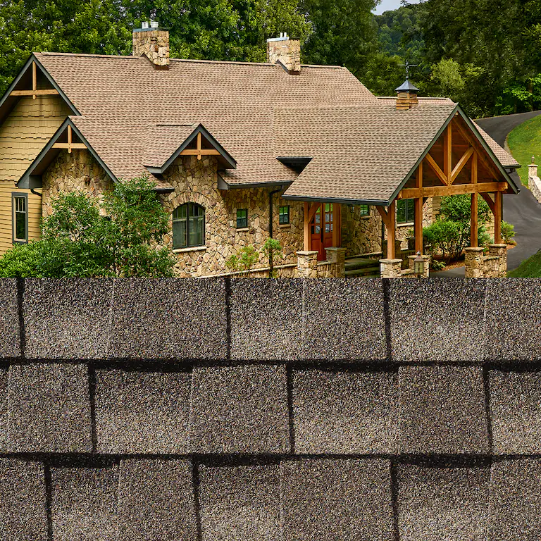 Stone house with tan shingled roof and wooden porch, seen behind a gray block wall and trees.
