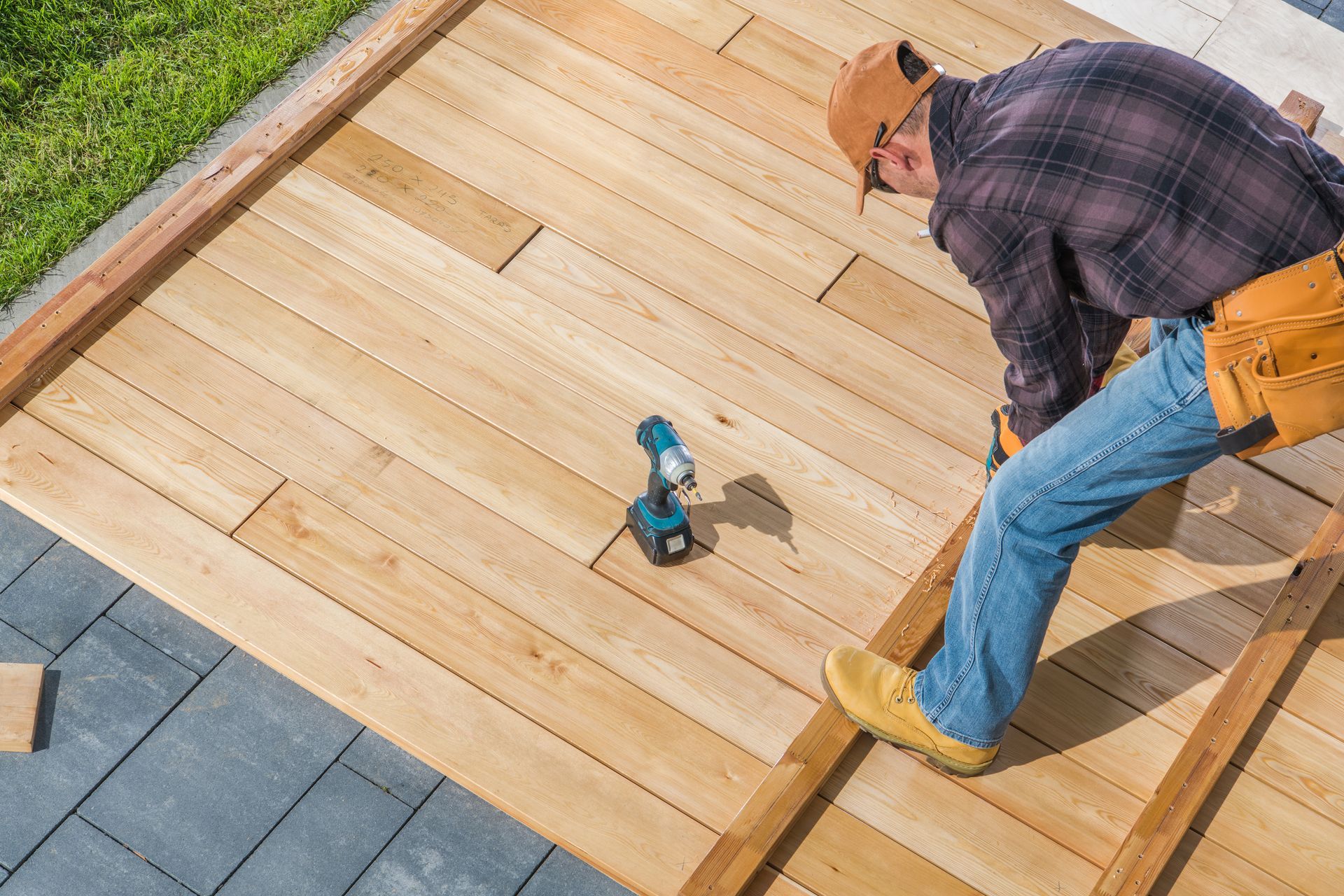Worker installing wooden deck boards with a drill on a backyard patio.