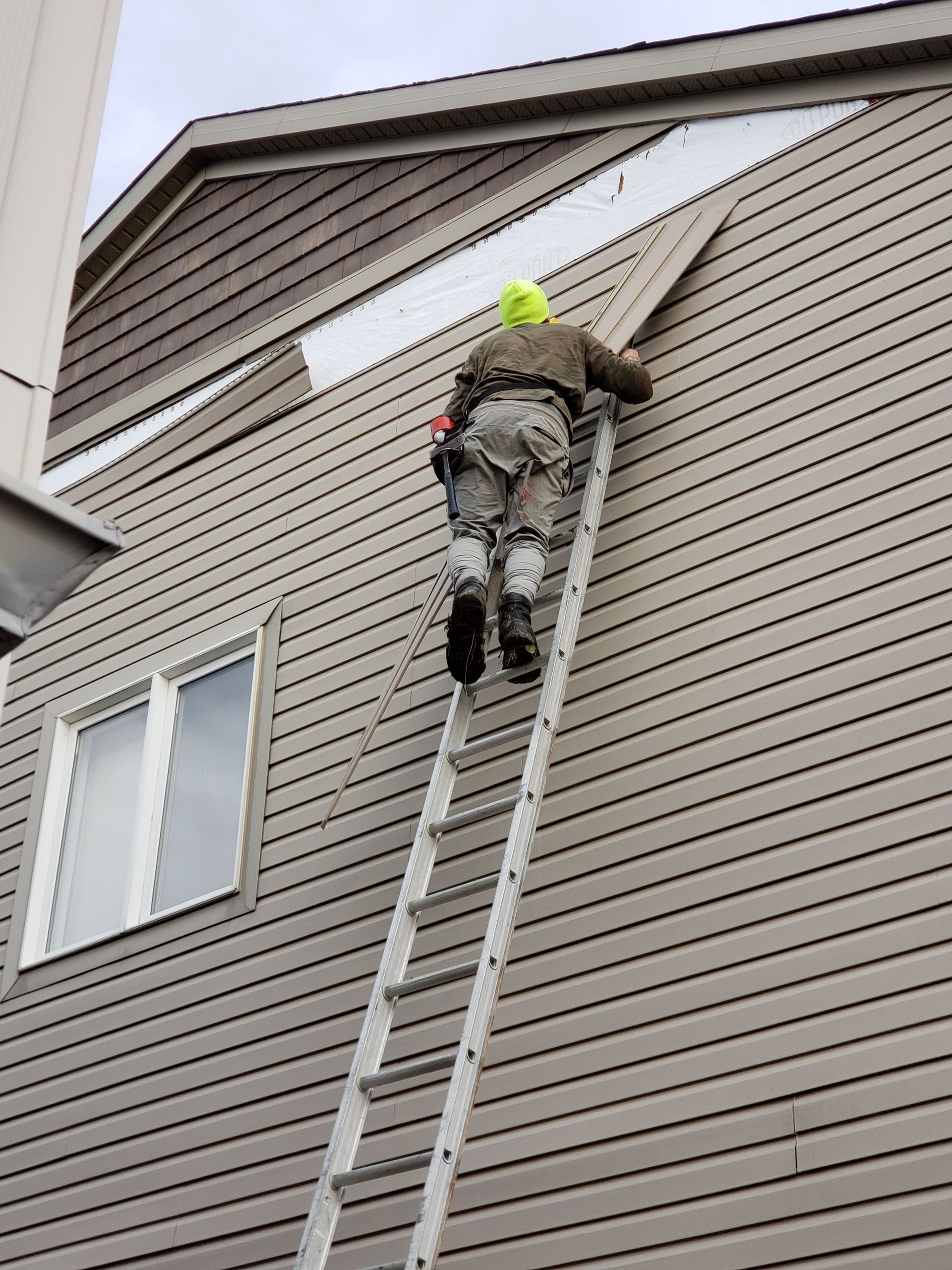 Worker on a ladder repairing house siding near the roofline, wearing a neon hat and safety harness.