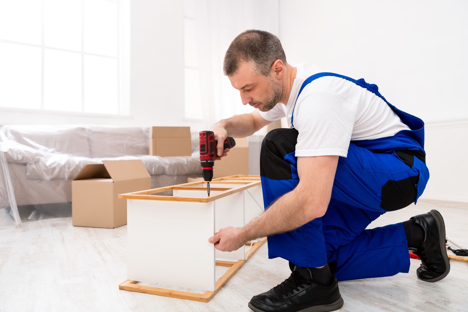 A person in blue overalls kneels, assembling furniture with a power drill indoors.