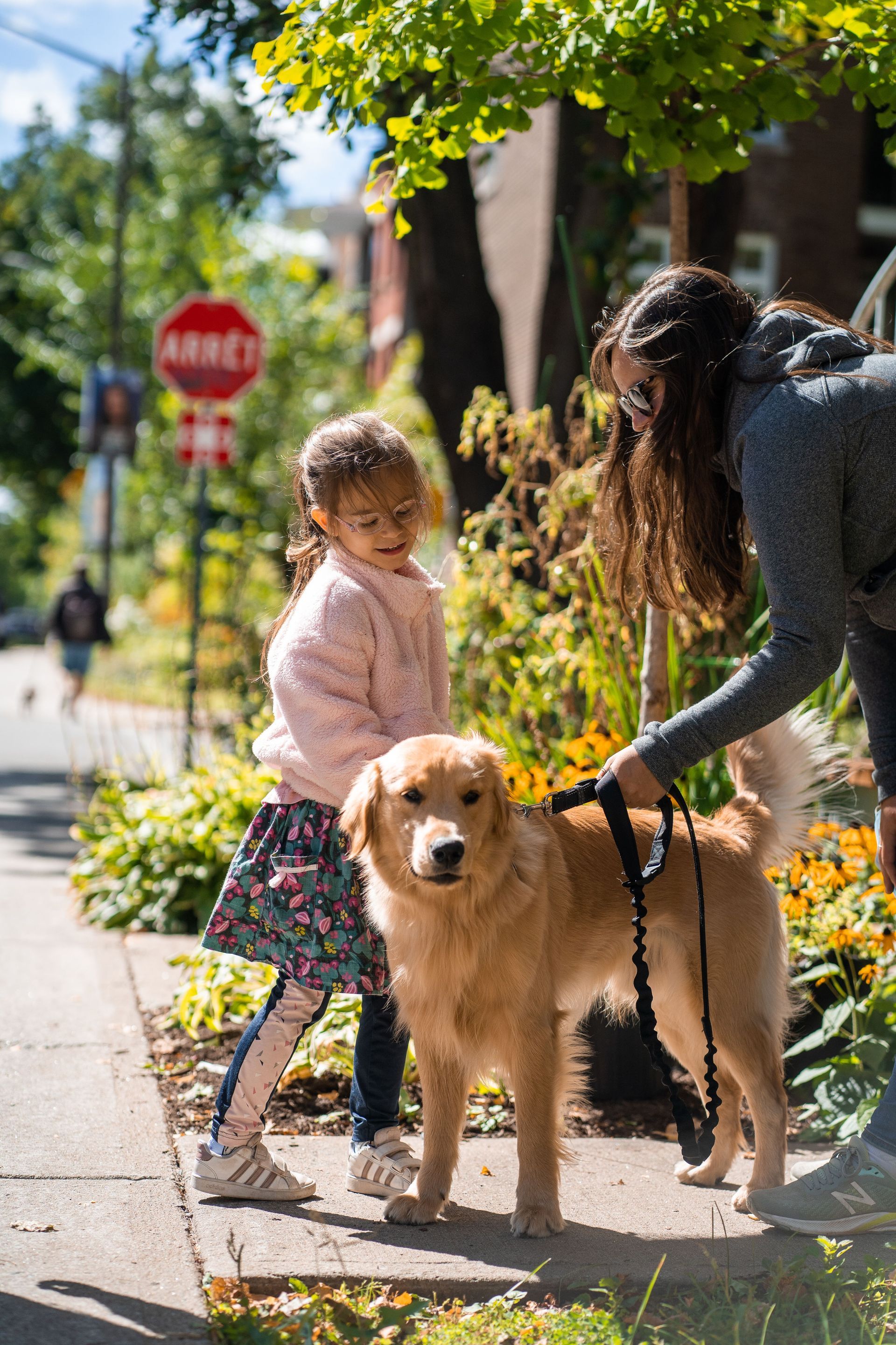 Woman and child pet a golden retriever on a sunny sidewalk with flowers nearby.