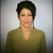Woman with dark hair smiles, wearing an olive green sweater and earrings, posing against a light background.
