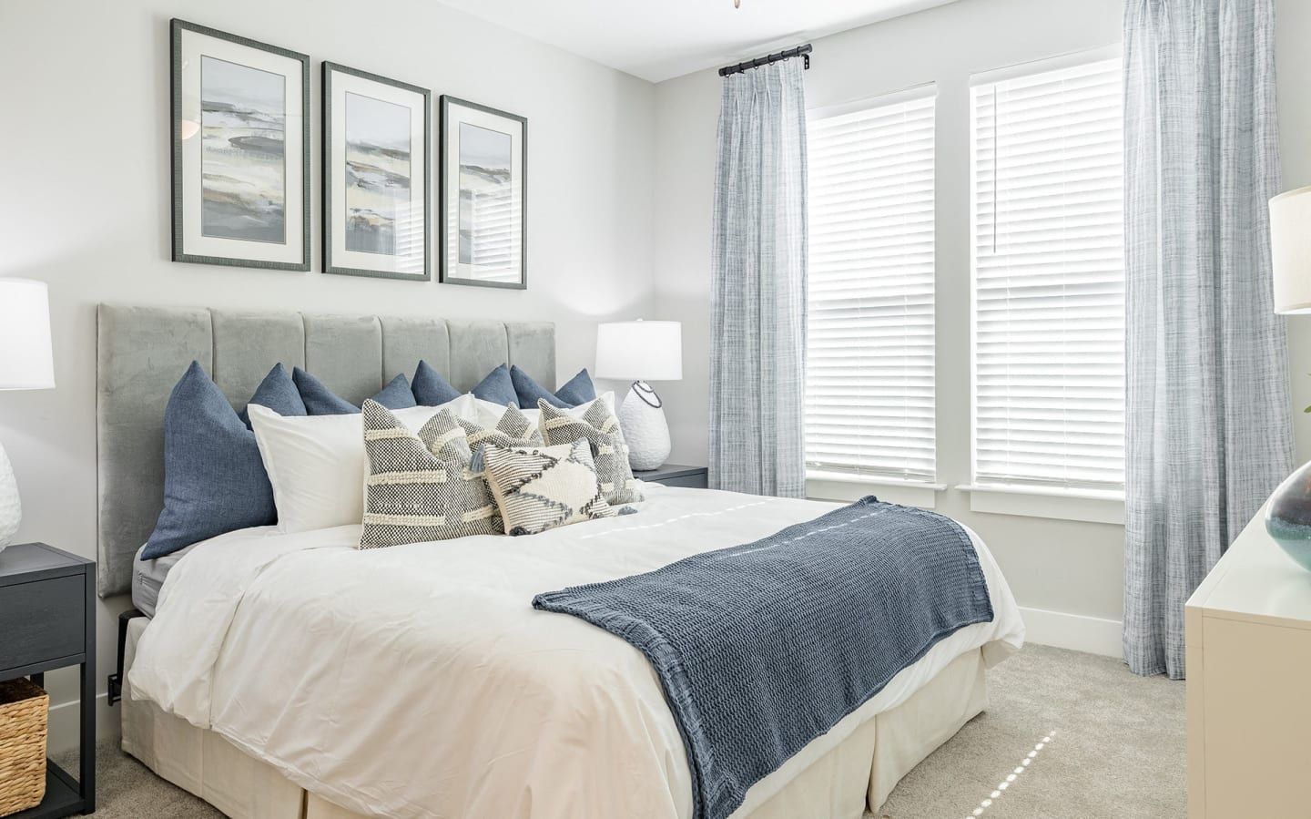 A bedroom with a bed , dresser , lamps , and three windows.