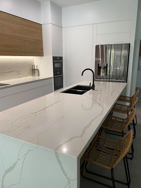 A white kitchen with a marble counter top and chairs under the bench 