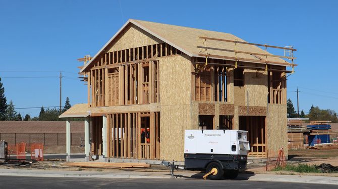 A two-story wood-framed house under construction, with a portable generator in the foreground on a sunny day.
