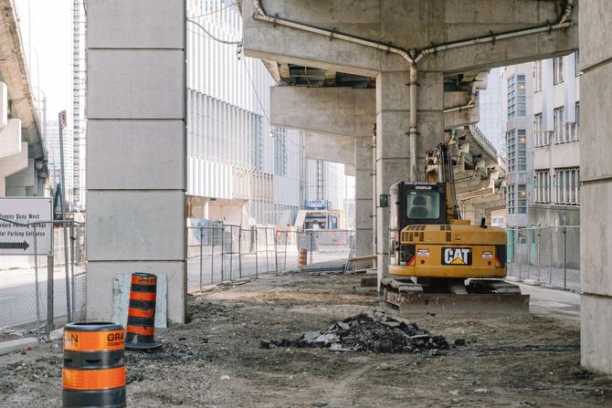 A yellow Caterpillar excavator sits on a gravel lot under elevated concrete highway structures with orange traffic cones.