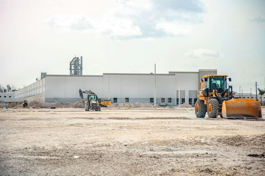 A yellow tractor stands in the foreground of a large, flat, dirt construction site with a long white warehouse behind it.