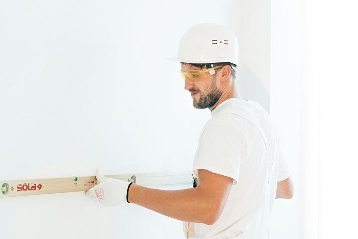 A person in a white hard hat and safety glasses holds a level against a white wall while performing construction work.