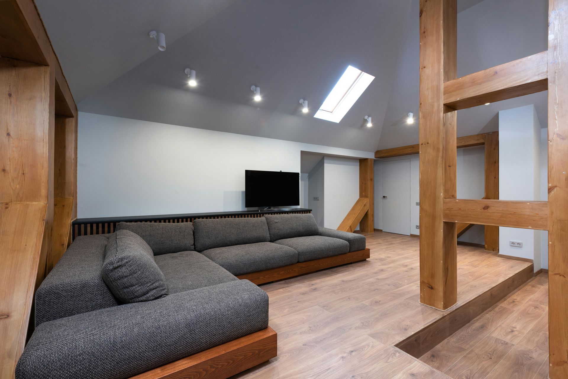 A spacious attic living room with a large, grey L-shaped sofa, light wood flooring, exposed timber beams, and a skylight.
