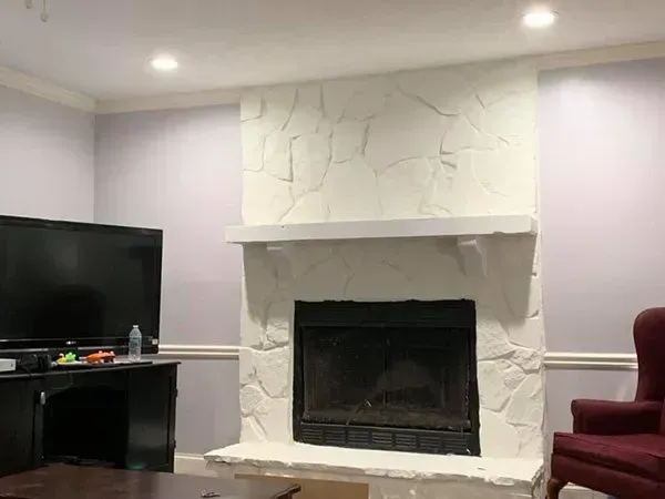 A room featuring a white stone fireplace as a focal point, a black TV console to the left, and a maroon armchair nearby.