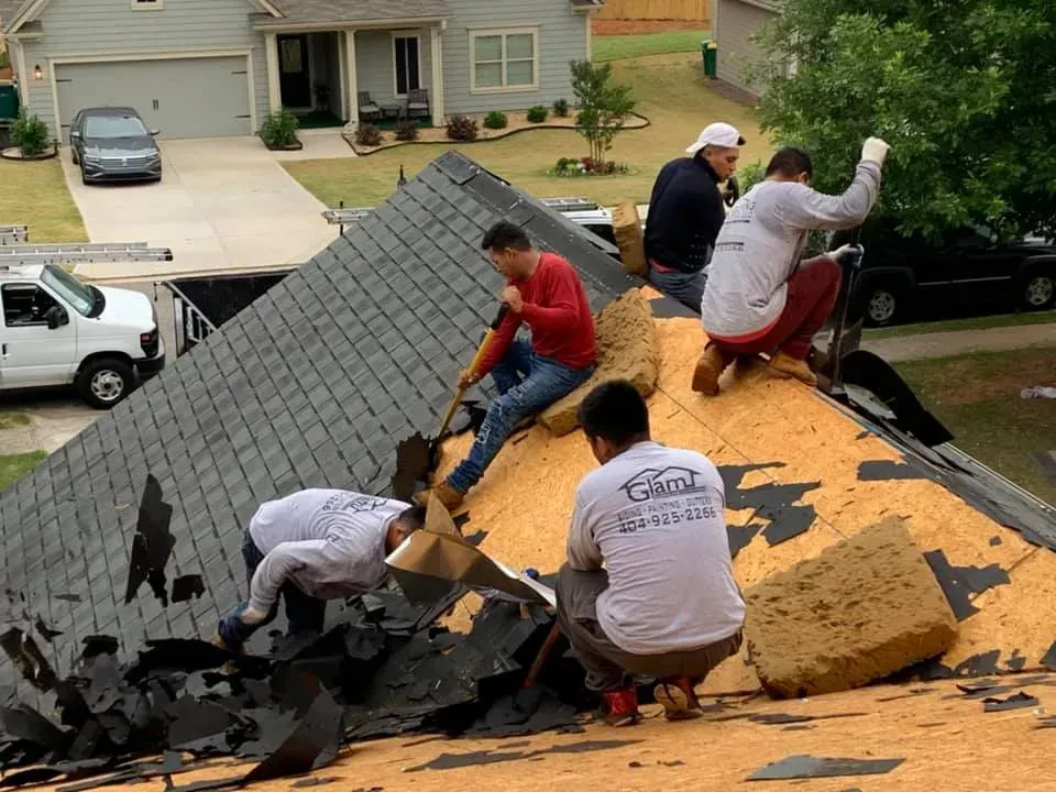 Four workers in work clothes stripping old shingles off a suburban house roof.