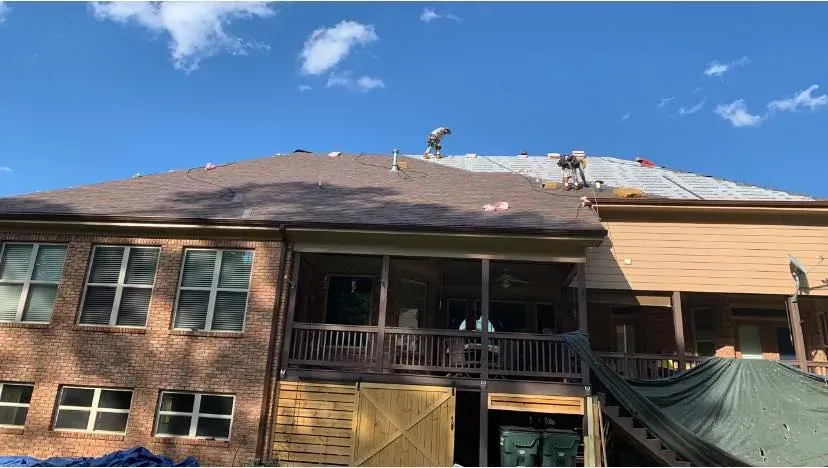 Workers replace shingles on a residential roof on a sunny day, with protective tarps placed below the structure.