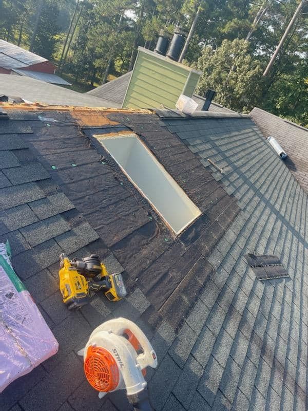 A partially shingled residential roof with a skylight opening, a yellow nail gun, and an orange blower tool.