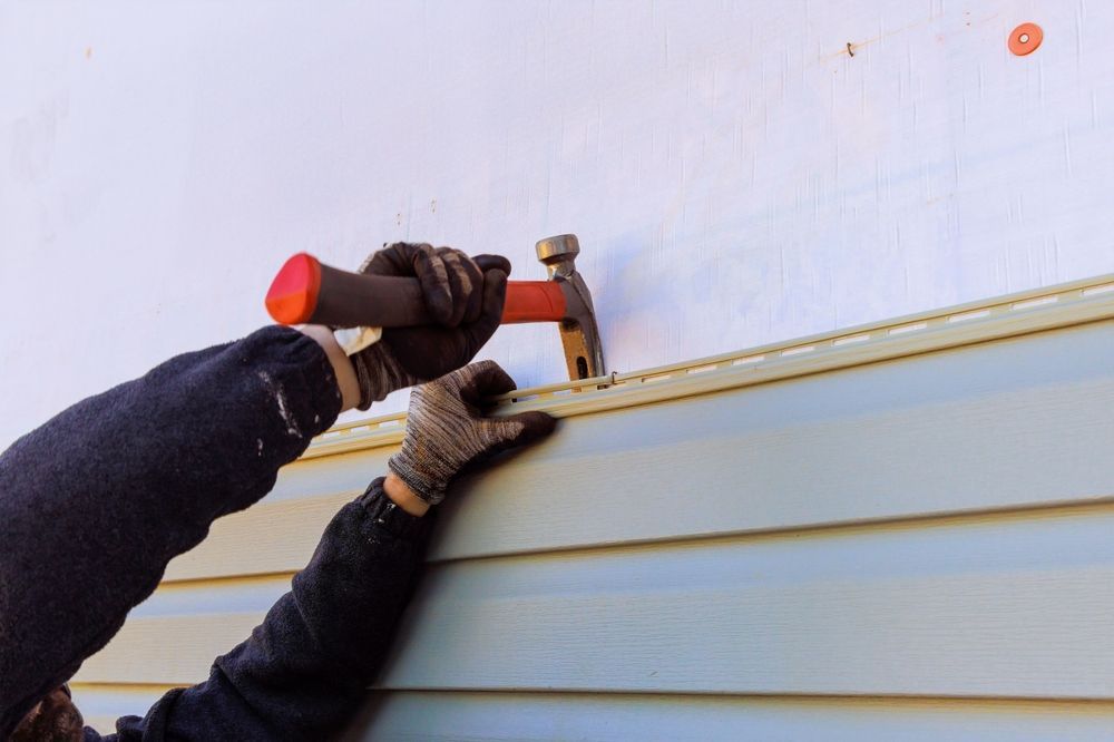 A person wearing gloves uses a hammer to install light-colored vinyl siding on an exterior wall.