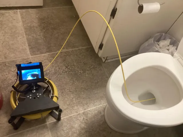 Robot with camera near a toilet, connected by a yellow cable, in a bathroom with tiled floor.