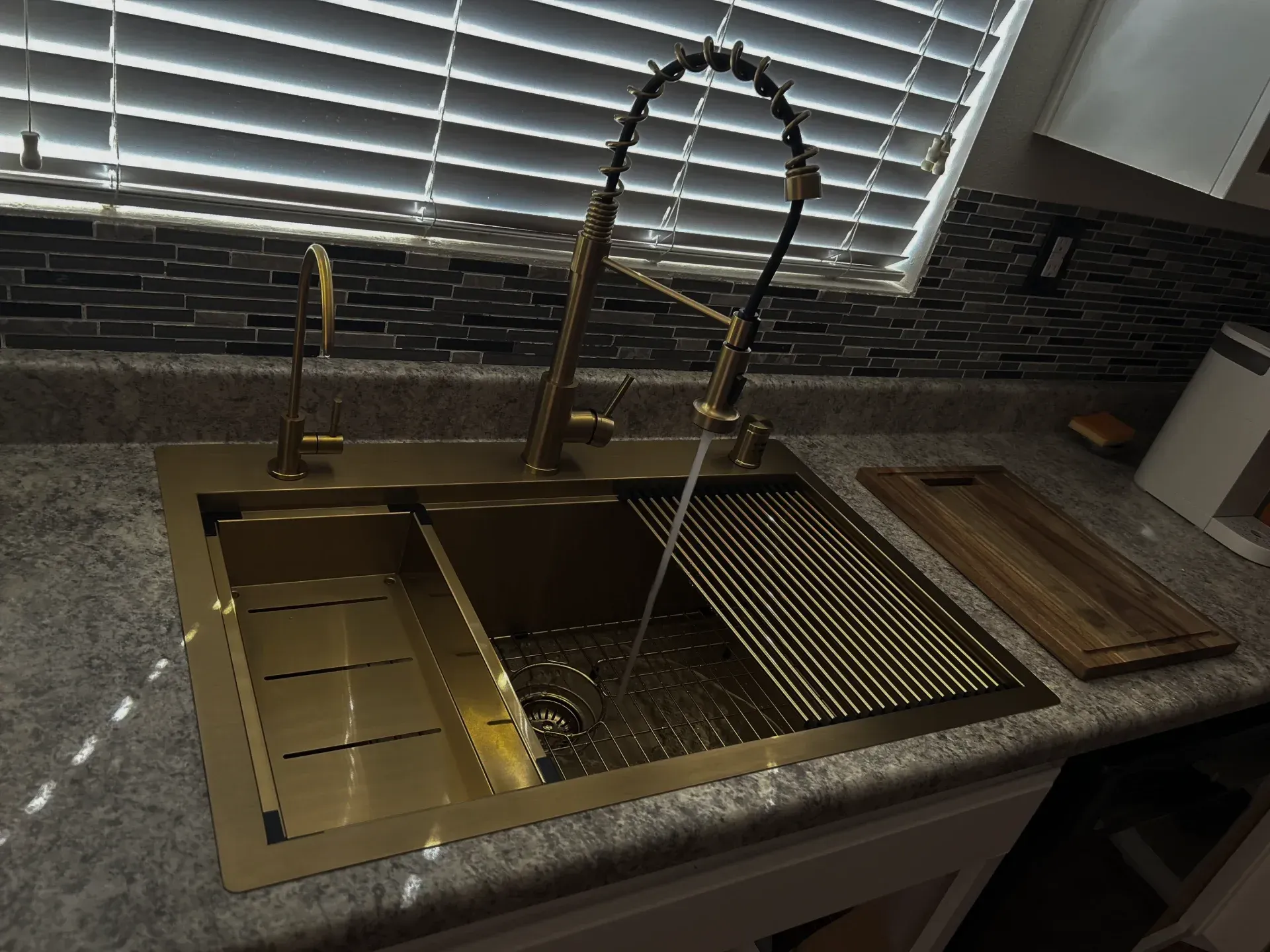 Kitchen sink with gold faucet, drying rack, and granite countertop by a window with blinds