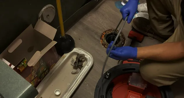 Person in blue gloves works on a drain pipe beside a sink and toilet in a bathroom.