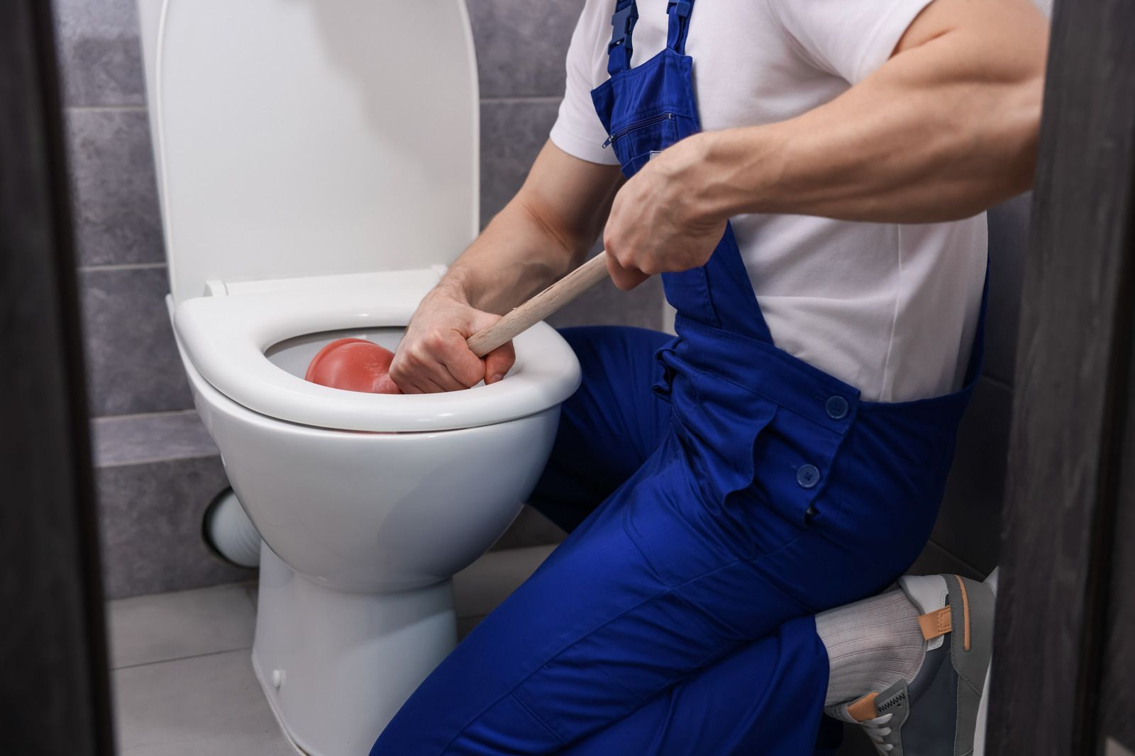 Person in blue overalls plunging a toilet with a red rubber cup in a bathroom