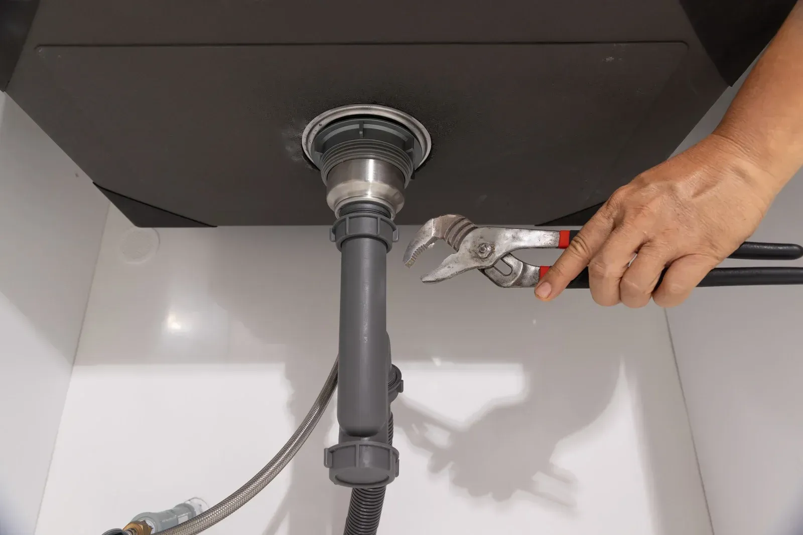 Hand tightening a sink drain pipe fitting under a black sink with pliers