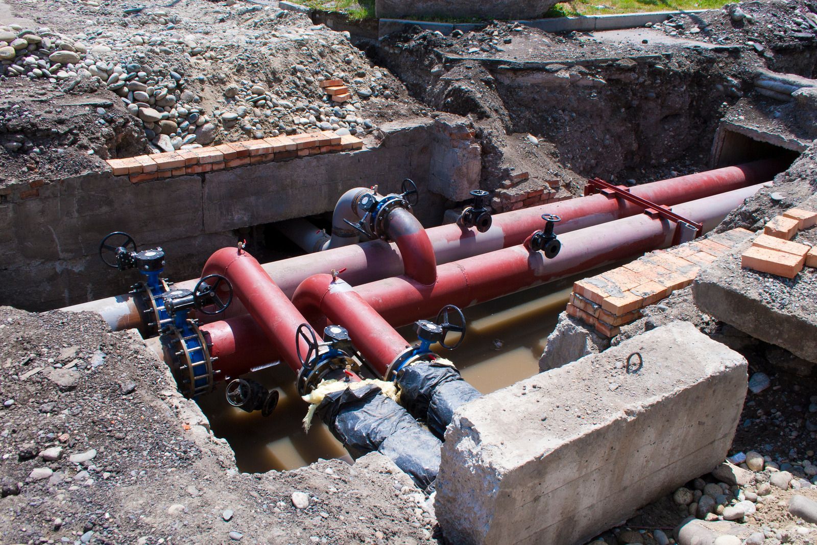 Red industrial pipes and valves crossing an excavated trench with muddy water and dirt piles