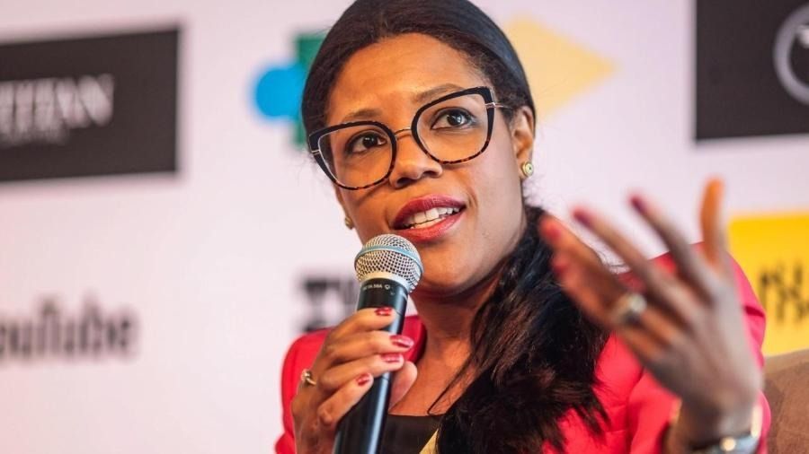 Woman speaking into a microphone, gesturing with her right hand. She wears glasses and a red jacket.