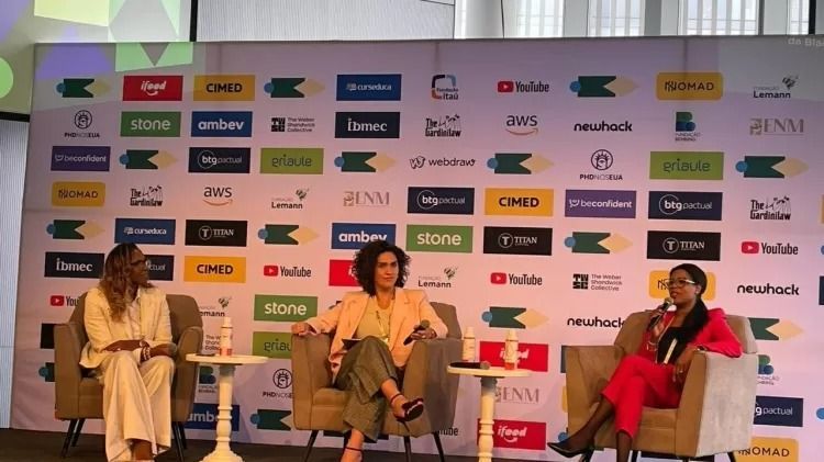 Three women seated on a stage, a backdrop with sponsor logos, talking at a conference.