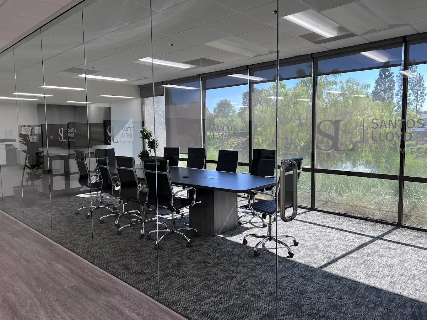 A modern glass-walled conference room with a long table, ergonomic chairs, and large windows looking out onto trees.