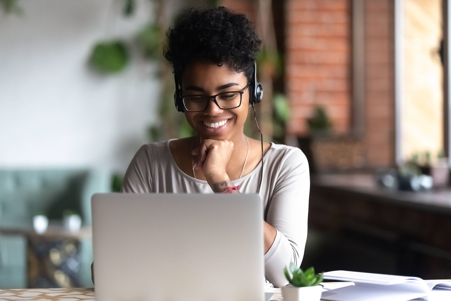 Woman with glasses and headphones smiling at a laptop, indoors near a window.