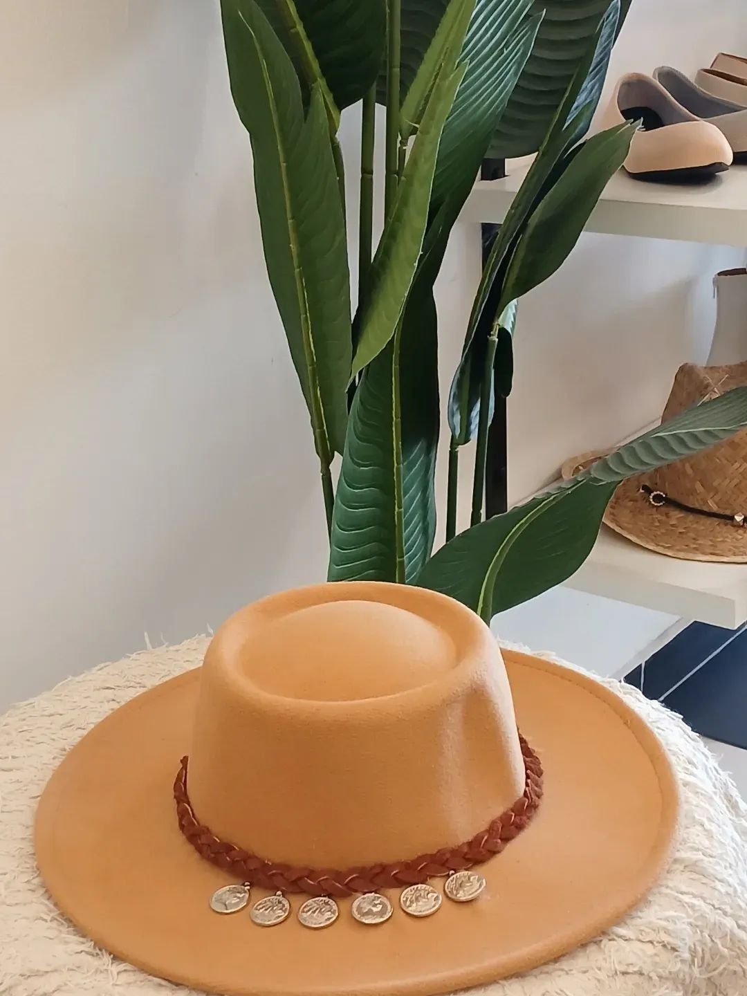 Un sombrero color canela con una banda de cuentas marrones sobre una mesa blanca, delante de hojas verdes y otros sombreros.