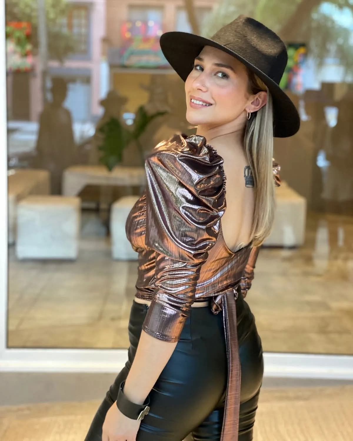 Mujer con top color bronce metalizado y sombrero negro posando en el interior, mirando por encima del hombro y sonriendo.