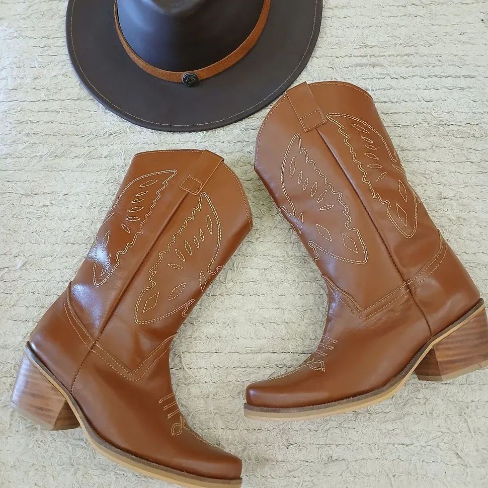 Botas vaqueras marrones con detalles de costura y un sombrero negro sobre un fondo claro.