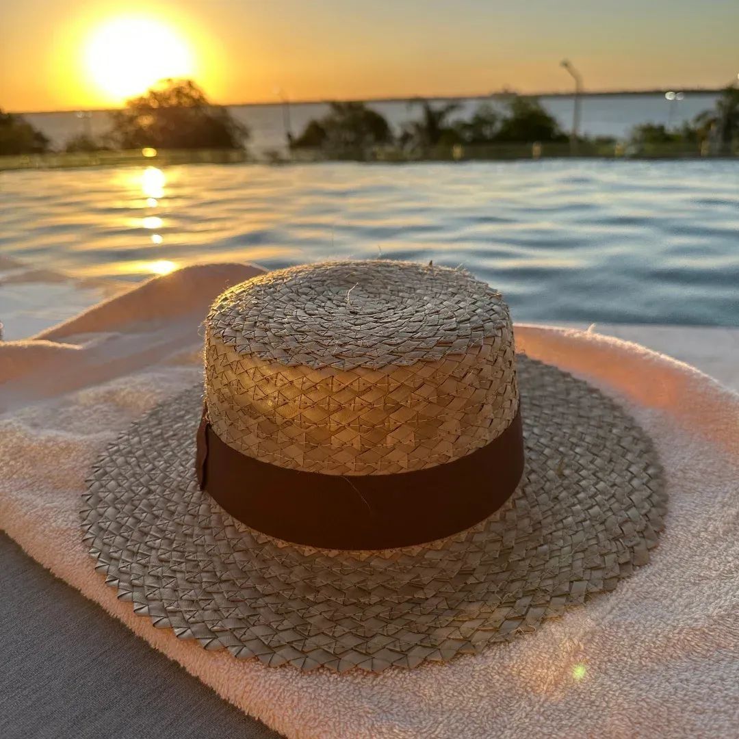 Sombrero de paja sobre una toalla junto a una piscina al atardecer, con agua y luz brillante de fondo.