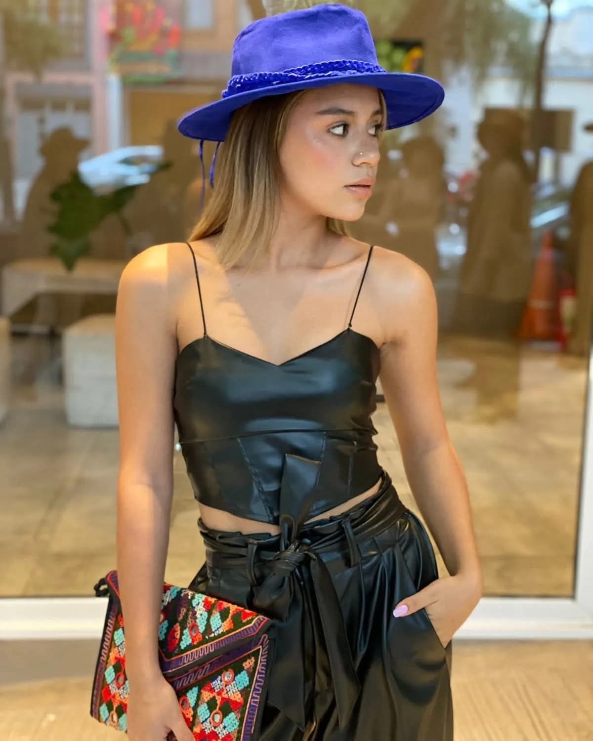 Mujer con sombrero azul y atuendo negro de pie en el interior de un edificio, con un bolso estampado de colores.