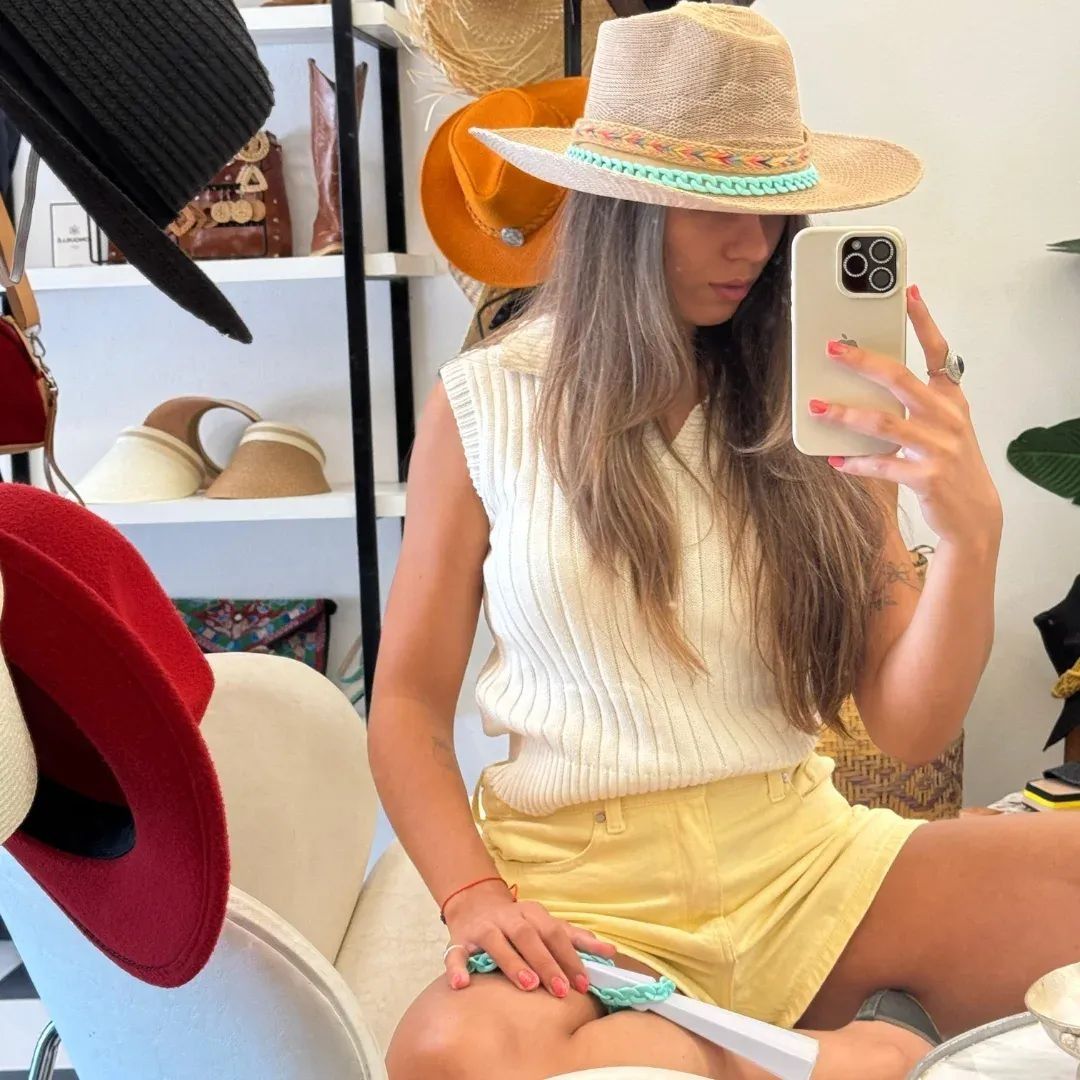 Mujer con sombrero de paja tomándose una selfie frente al espejo mientras está sentada en una sombrerería luminosa.