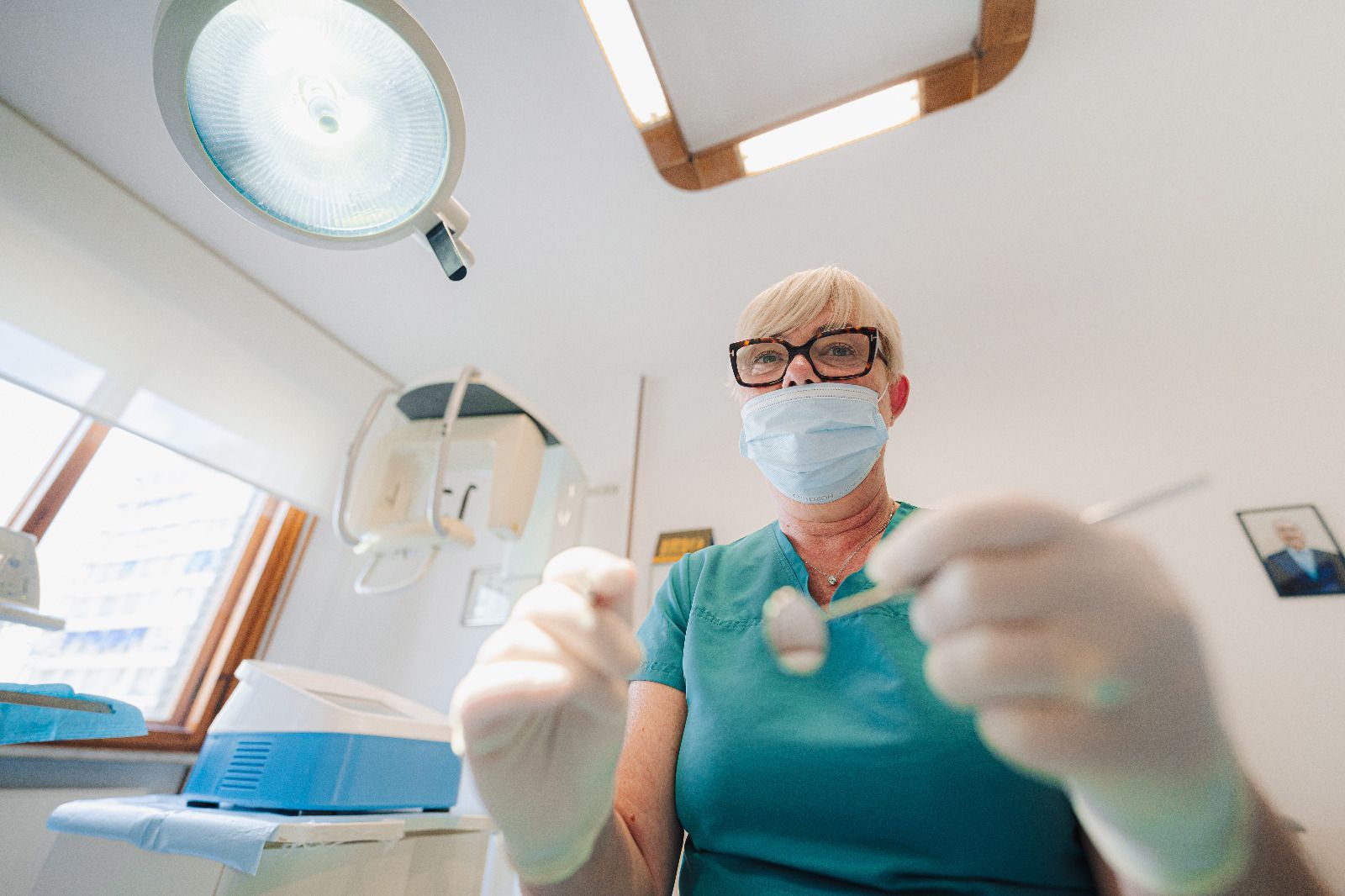 Un dentista con mascherina e guanti, in una luminosa clinica odontoiatrica, tiene uno strumento dentale rivolto verso la telecamera.