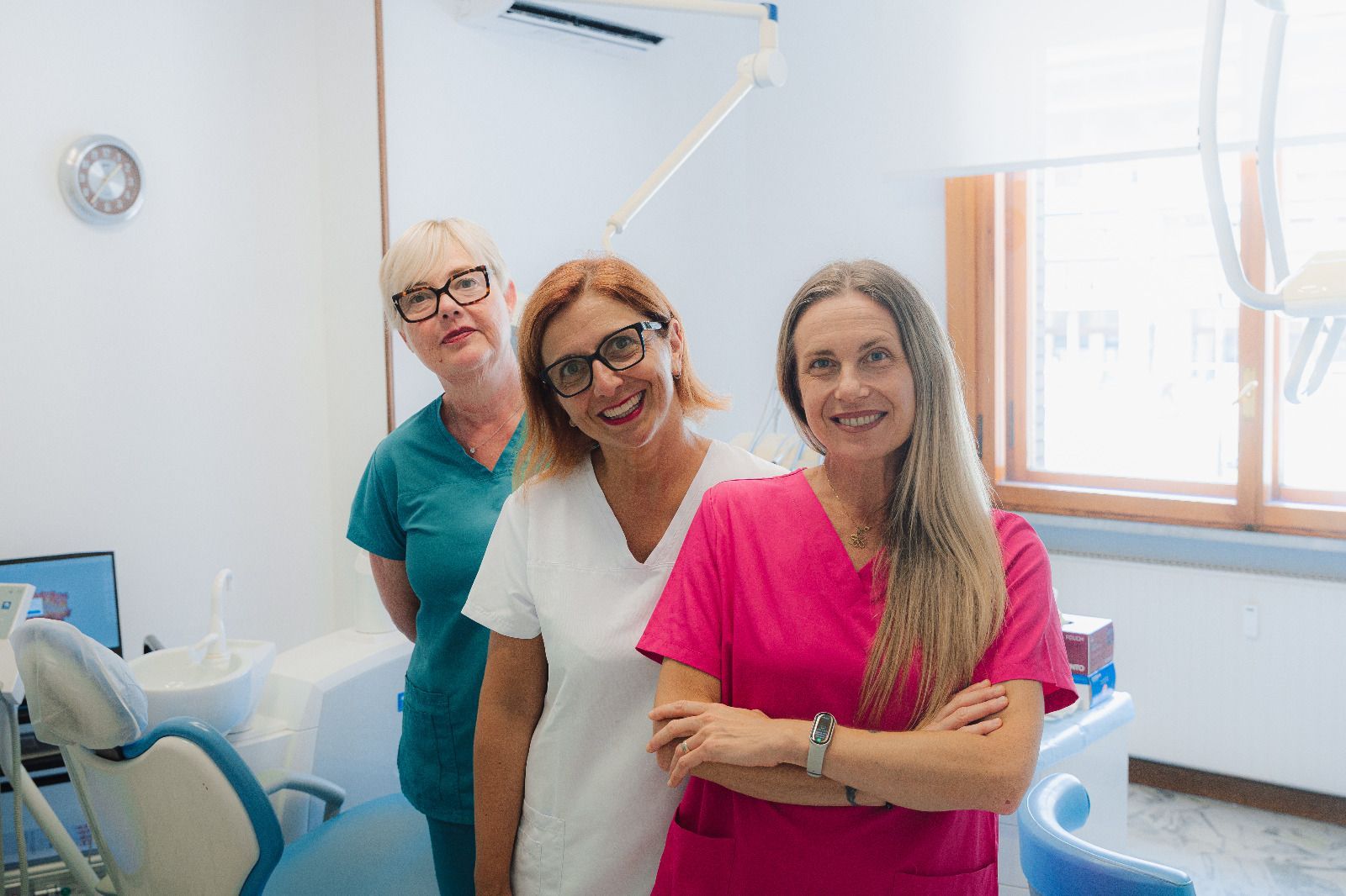 Tre membri sorridenti dello staff odontoiatrico in una clinica luminosa, in piedi accanto a una poltrona odontoiatrica.