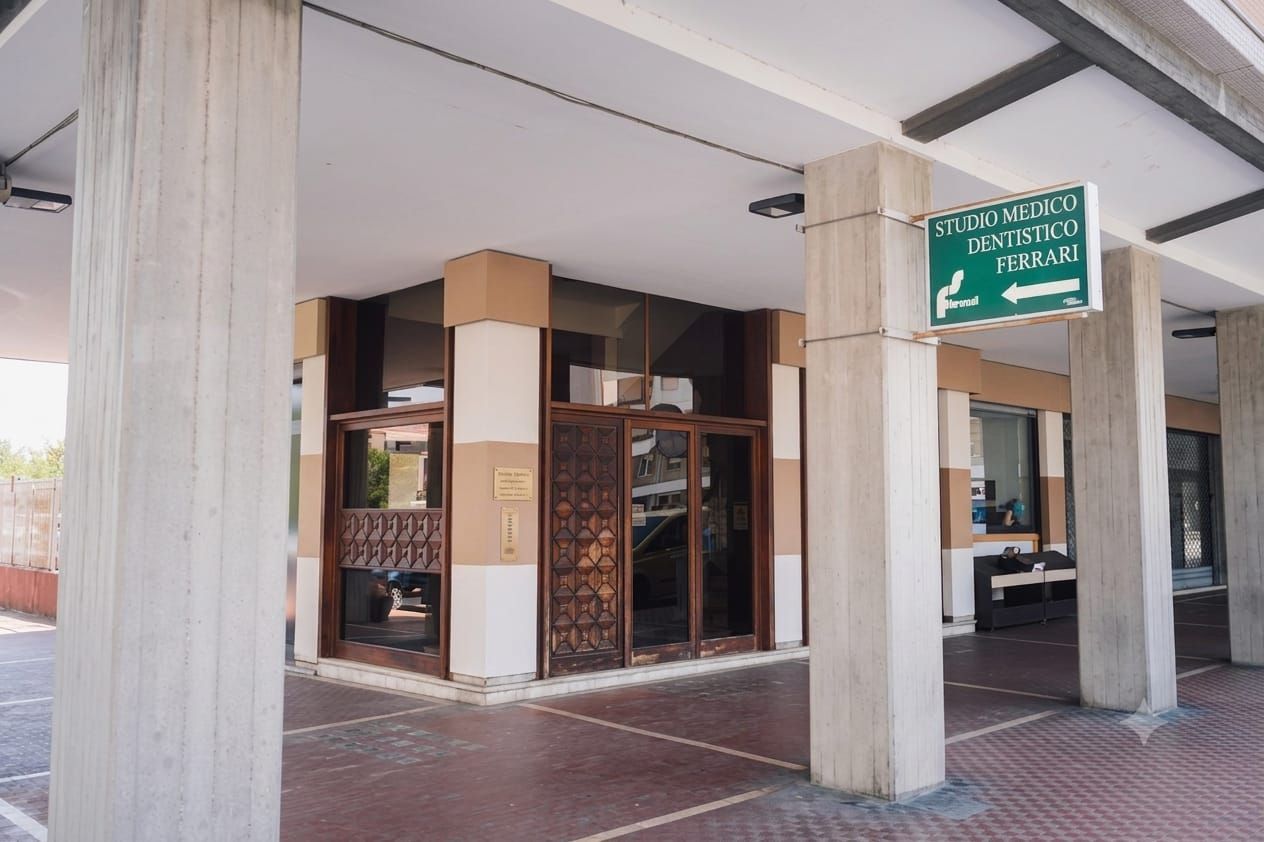 Ingresso coperto dell'edificio con colonne in cemento, porte a vetri e un'insegna verde sospesa.