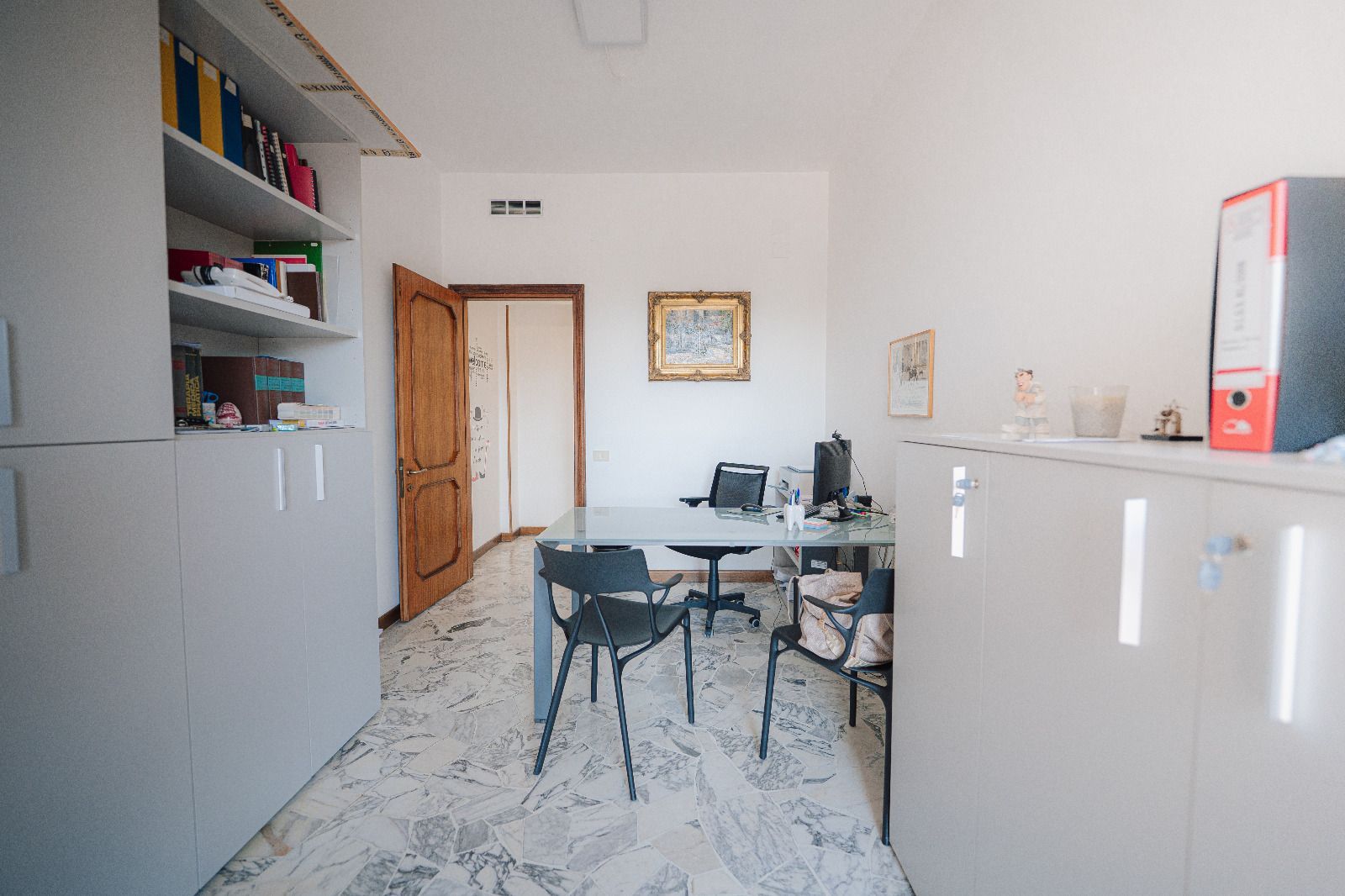 Piccolo ufficio con scrivanie, sedie, armadietti e una porta di legno in una stanza luminosa.