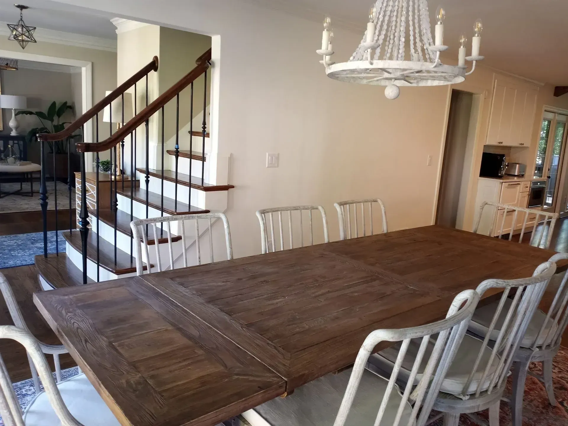 Dining room with a large wooden table, white chairs, staircase, and chandelier in a bright home interior
